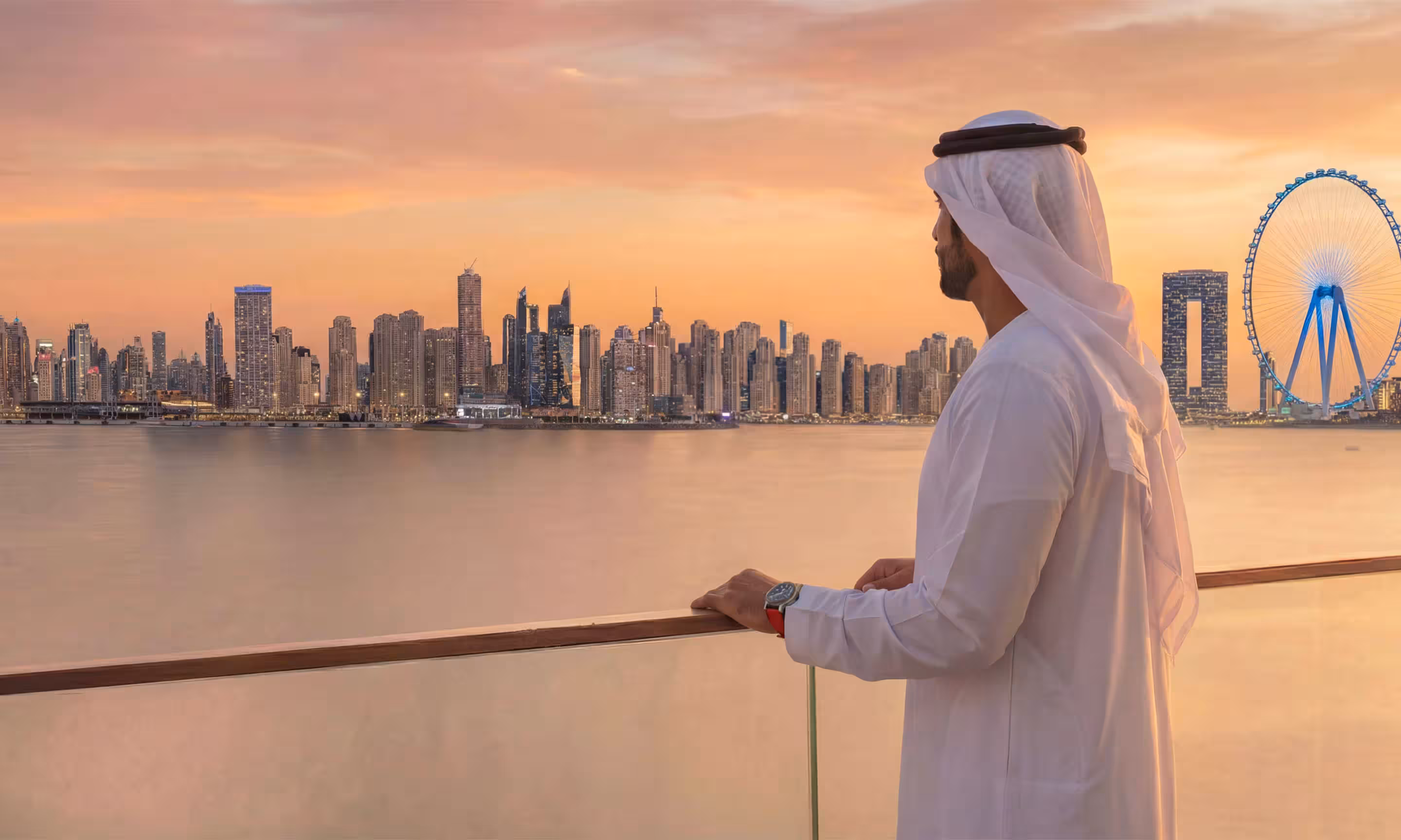 Man in traditional Emirati clothing looking at Dubai skyline at sunset over calm water.